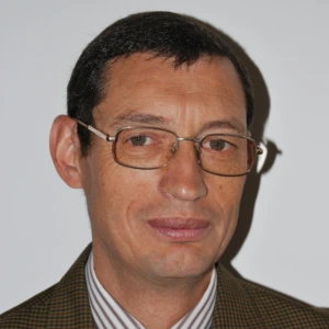 Vidutinio amžiaus vyras trumpais tamsiais plaukais ir akiniais su plonais metaliniais rėmeliais. Jis dėvi smulkiai languotą ruda ir žalsvą &scaron;varką bei dryžuotus mar&scaron;kinius. Vyras žiūri į kamerą ir lengvai &scaron;ypsosi &scaron;viesame fone.