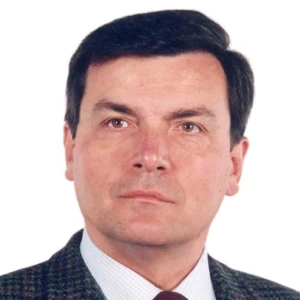 Vyras trumpais, tvarkingai su&scaron;ukuotais tamsiais plaukais, žiūrintis tiesiai į kamerą. Jis dėvi tamsų languotą &scaron;varką, &scaron;viesius mar&scaron;kinius ir tamsiai raudoną kaklarai&scaron;tį. Nuotraukos fonas yra vientisai baltas.