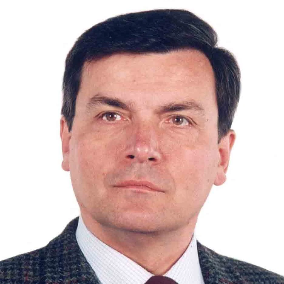 Vyras trumpais, tvarkingai sušukuotais tamsiais plaukais, žiūrintis tiesiai į kamerą. Jis dėvi tamsų languotą švarką, šviesius marškinius ir tamsiai raudoną kaklaraištį. Nuotraukos fonas yra vientisai baltas.