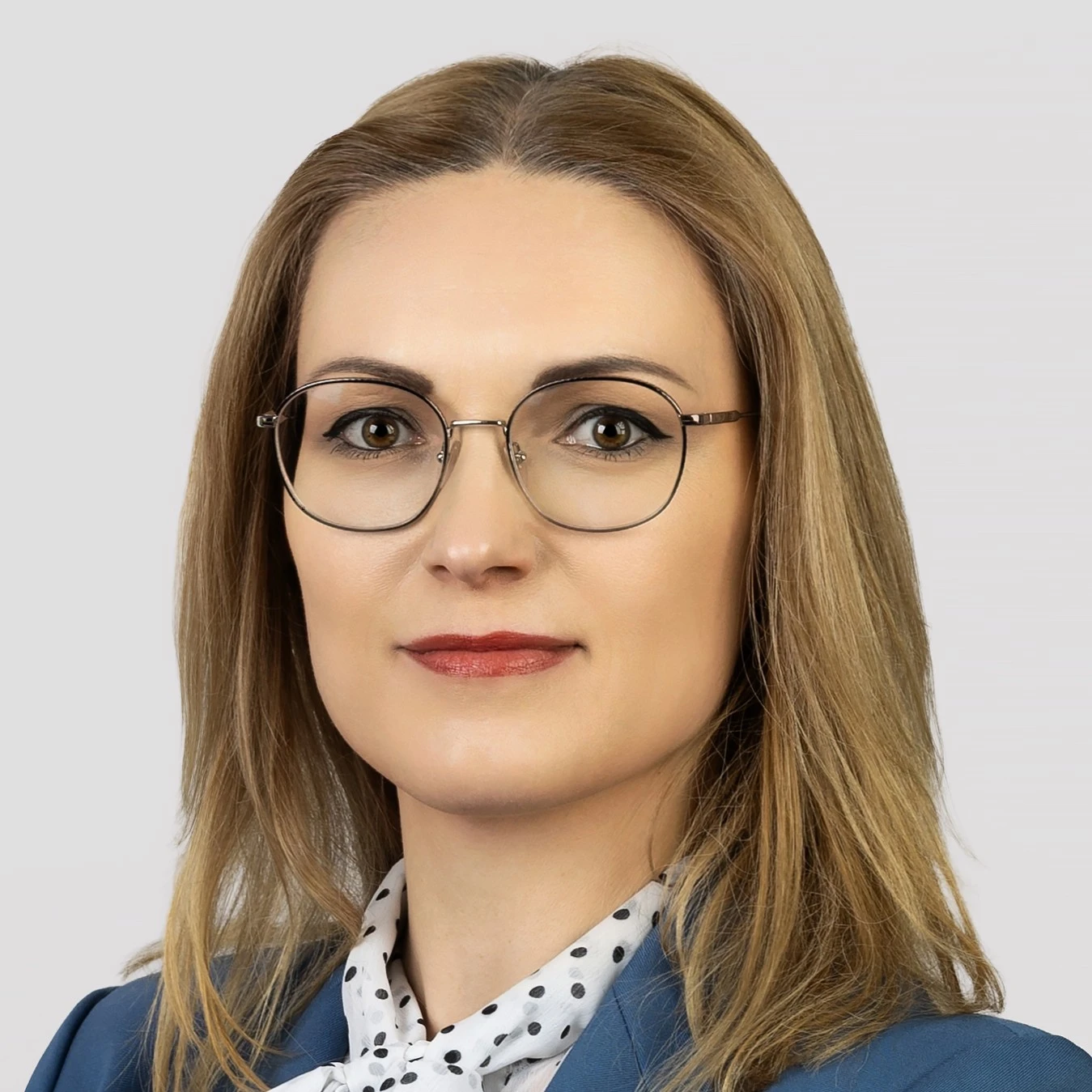 Moteris ilgais, tiesiais šviesiais plaukais ir apvaliais akiniais plonais rėmeliais. Ji lengvai šypsosi, dėvi melsvą švarką ir baltą palaidinę su juodais taškeliais. Fonas neutralus, šviesiai pilkas.