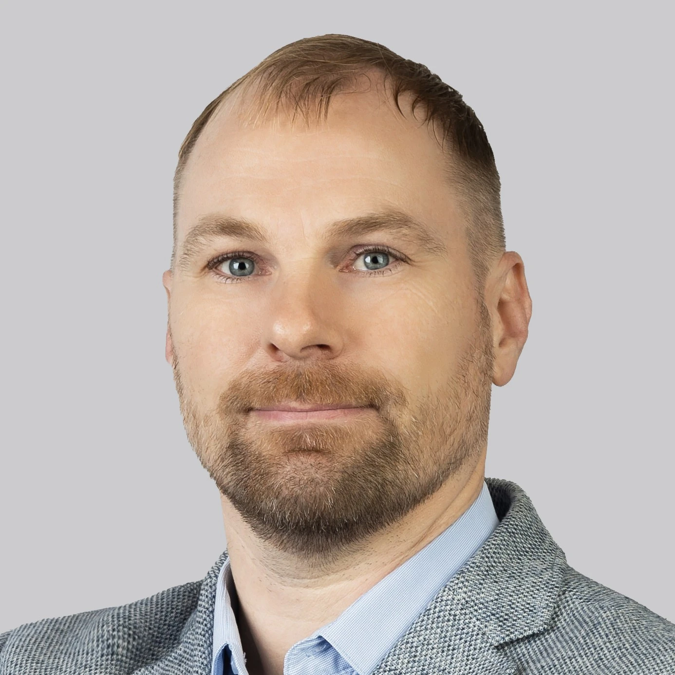 Vyras trumpais šviesiais plaukais ir tvarkinga ruda barzda bei ūsais. Jis dėvi pilką languotą švarką ir šviesiai mėlynus marškinius. Nuotraukos fonas yra neutralus ir pilkas.